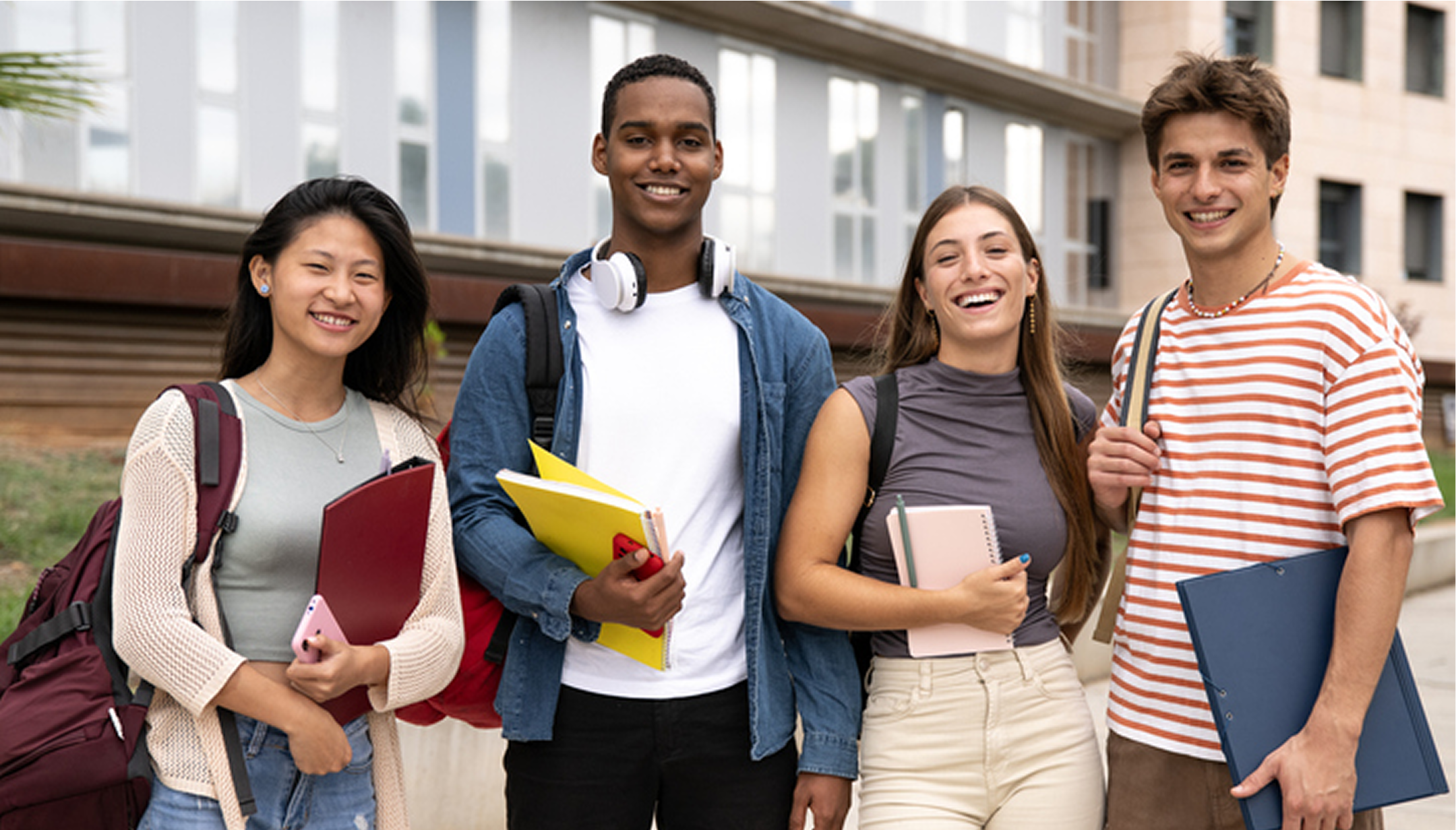 Four students smiling with books outside building.